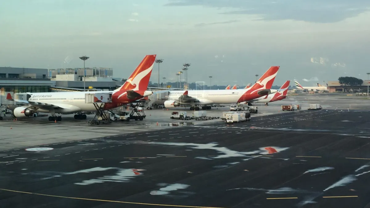 Three Qantas aircraft parked at their gates at dusk, seen through terminal glass, red kangaroo tail fins lined up along the pier