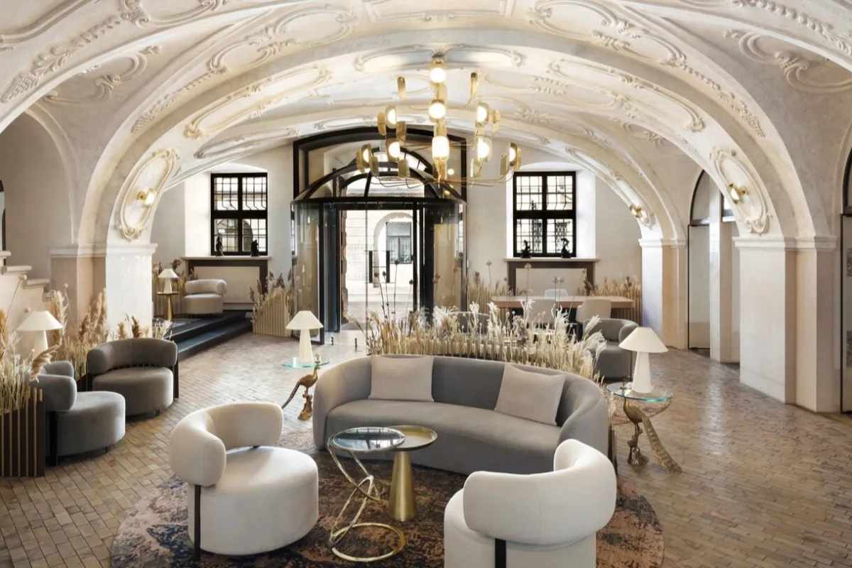 A bright hotel lobby with curved contemporary sofas and gold pendant lights beneath an ornate vaulted baroque ceiling