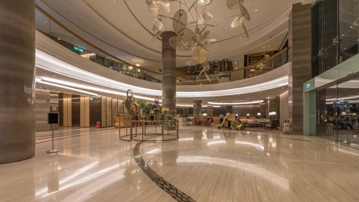 A bright, double-height hotel lobby with polished stone floors, marble columns and a curved mezzanine above the reception area