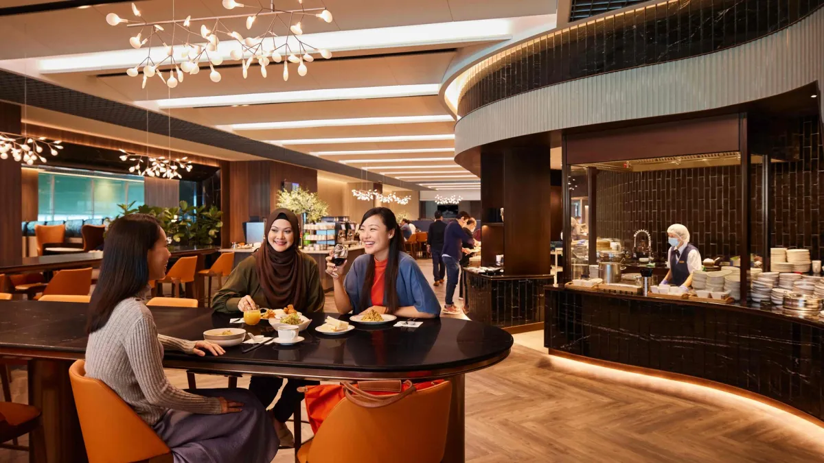 Three travellers dining together at a round table in the SilverKris Lounge dining hall at Singapore Changi Airport, with the open kitchen counter behind them